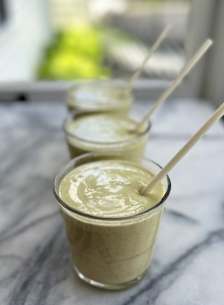 Three glasses with straws and mango protein smoothies on marble table in front of window.