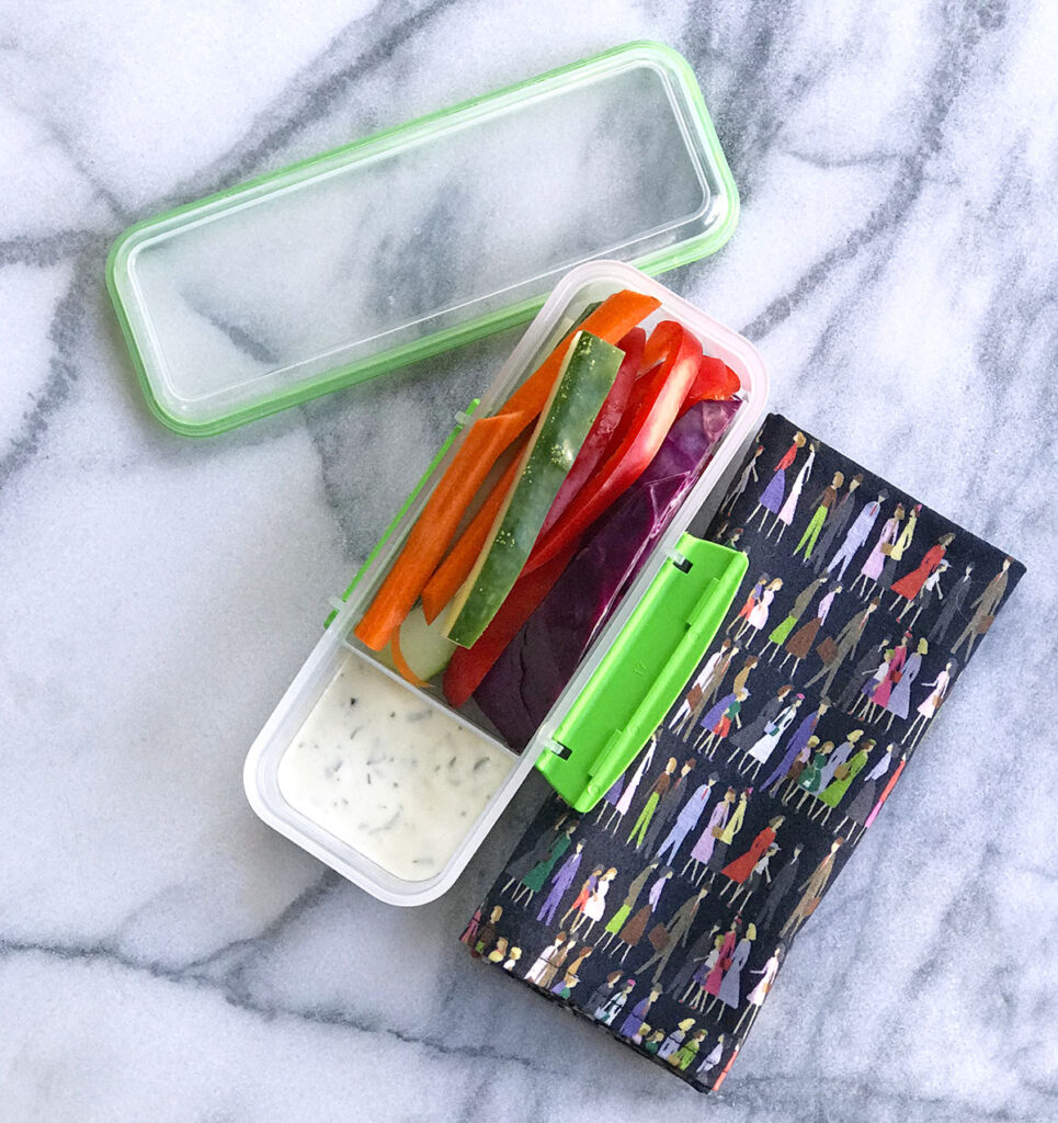 Plastic container of red pepper strips and cucumber strips with a compartment of ranch dip open on a counter next to a napkins with kids and adults in colorful clothes on it.