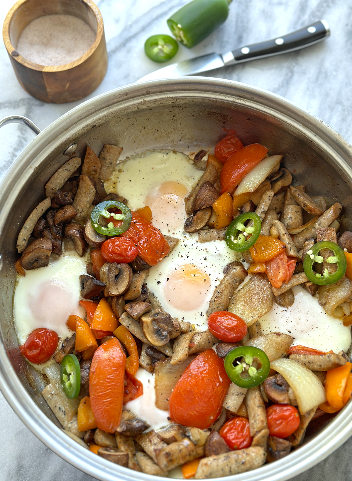 A large skillet has sausages, fried onions, tomatoes, mushrooms, orange peppers, and jalapenos with five eggs nestled in holes, all sitting with a salt container, knife, and raw jalapeno next to it on a marble surface. 