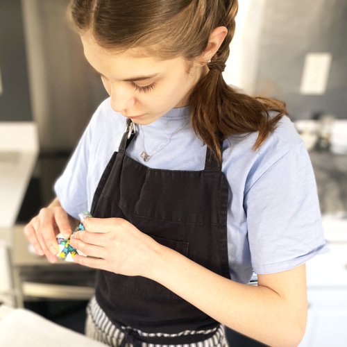 A girl with braids, black apron, and blue t-shirt wrapping washi tape around a hard-boiled egg as she learns how to color eggs using food dye for Easter.