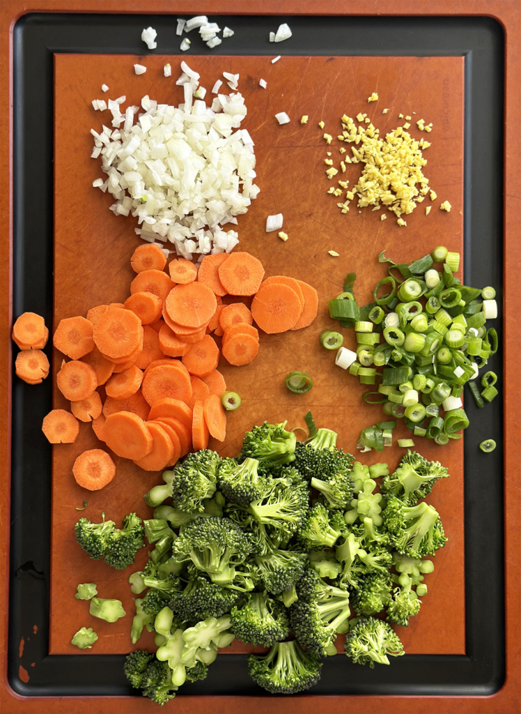 Brown Epicurean cutting board with black juice rim holding cut broccoli, carrots, onions, ginger and scallions. 