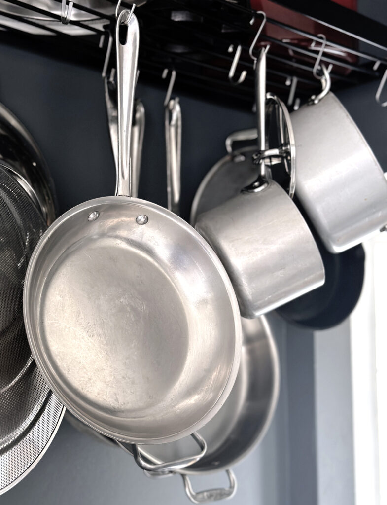 Stainless steel all-clad skillets handing from a pot rack in front of a blue wall.
