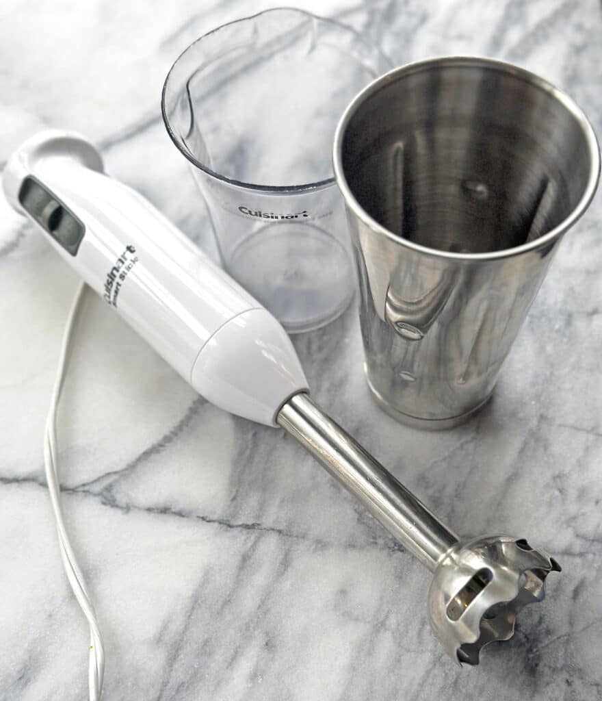 White Cuisinart Immersion blender with metal cup and clear plastic measuring cup for blending sitting on a marble counter. 