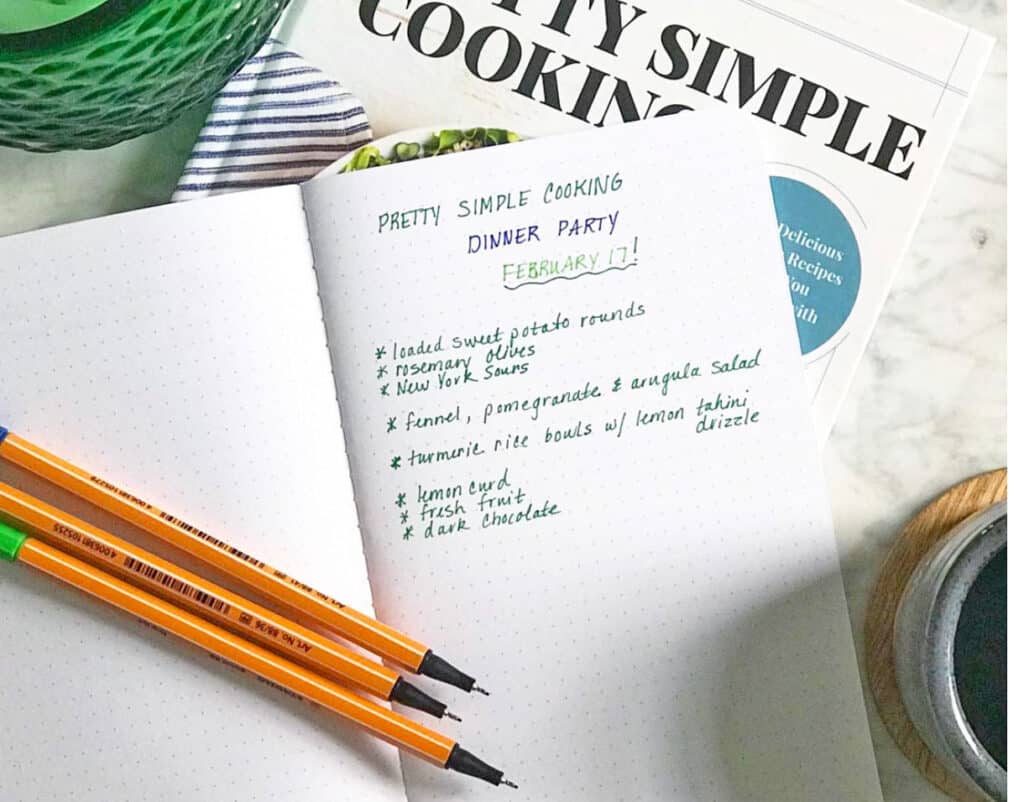 cook book with a notebeook on top with a menu for a dinner party with three pens sitting next to books