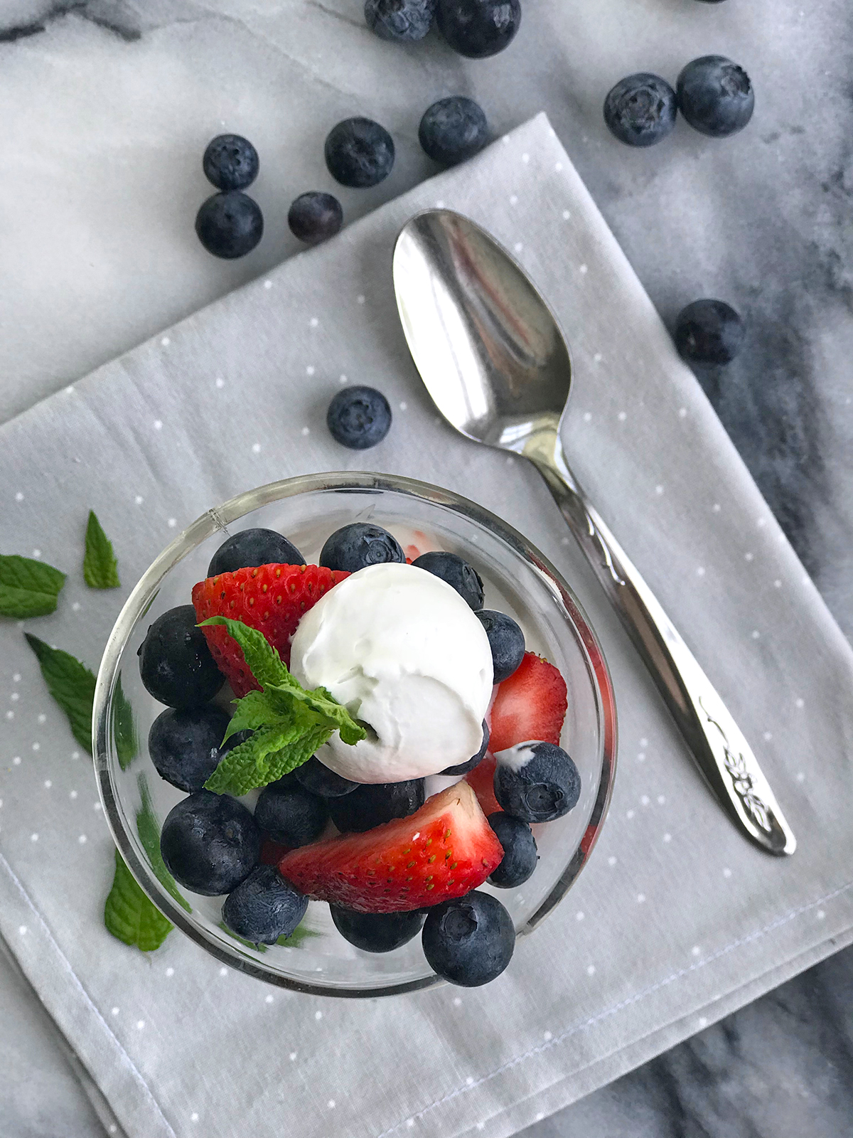 Glass dish with blueberries and strawberries with whippe coconut cream and a mint leaf on a gray napkin and spoon on marble.