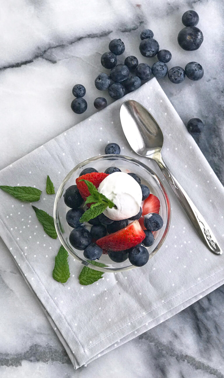 Glass dish with blueberries and strawberries with whippe coconut cream and a mint leaf on a gray napkin and spoon on marble.