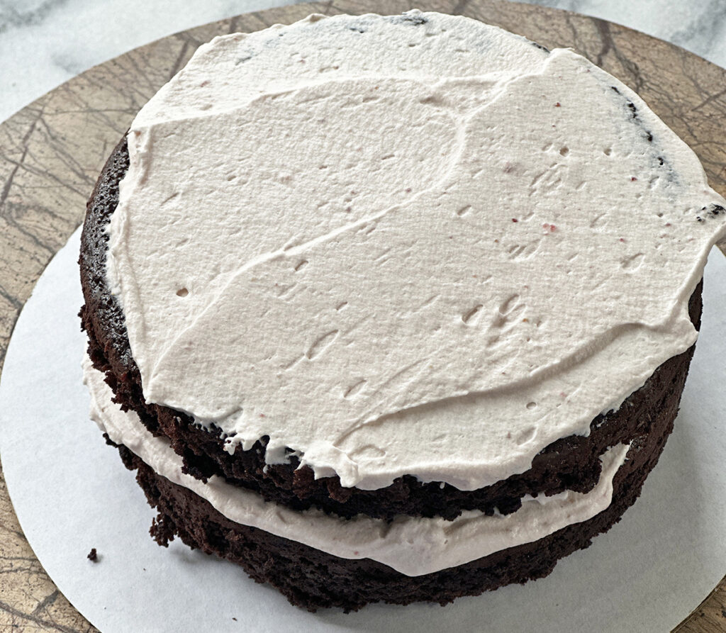 Two layers of chocolate cake with whipped cream frosting between and top sitting on a white cardboard cake round on a brown plate over marble surface. 