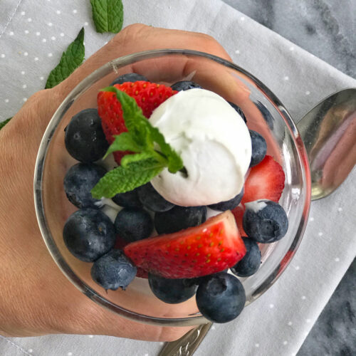 Hand holding clear glass parfait of fresh berries and dollop of coconut cream and mint on top.