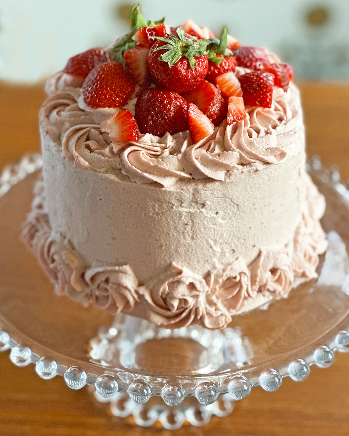 Small round layered cake with pink whipped cream frosting with swirls on top and fresh berries on top all sitting on glass cake pedestal on a wood table. 