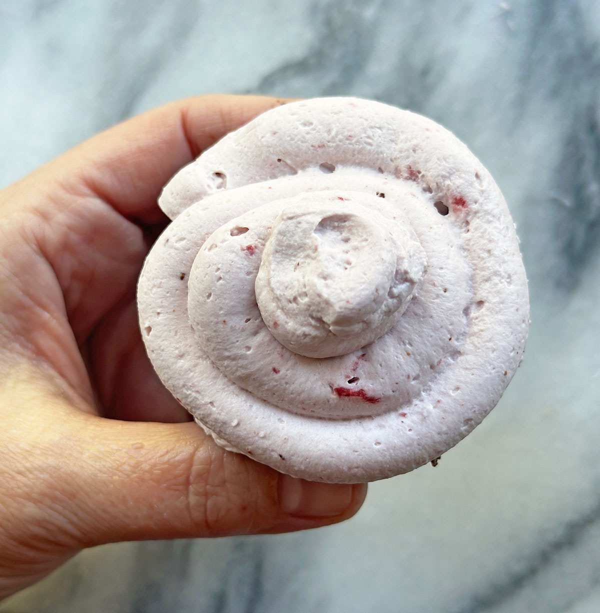 left hand holding a chocolate cupcake covered with swirl of strawberry pink coconut cream frosting