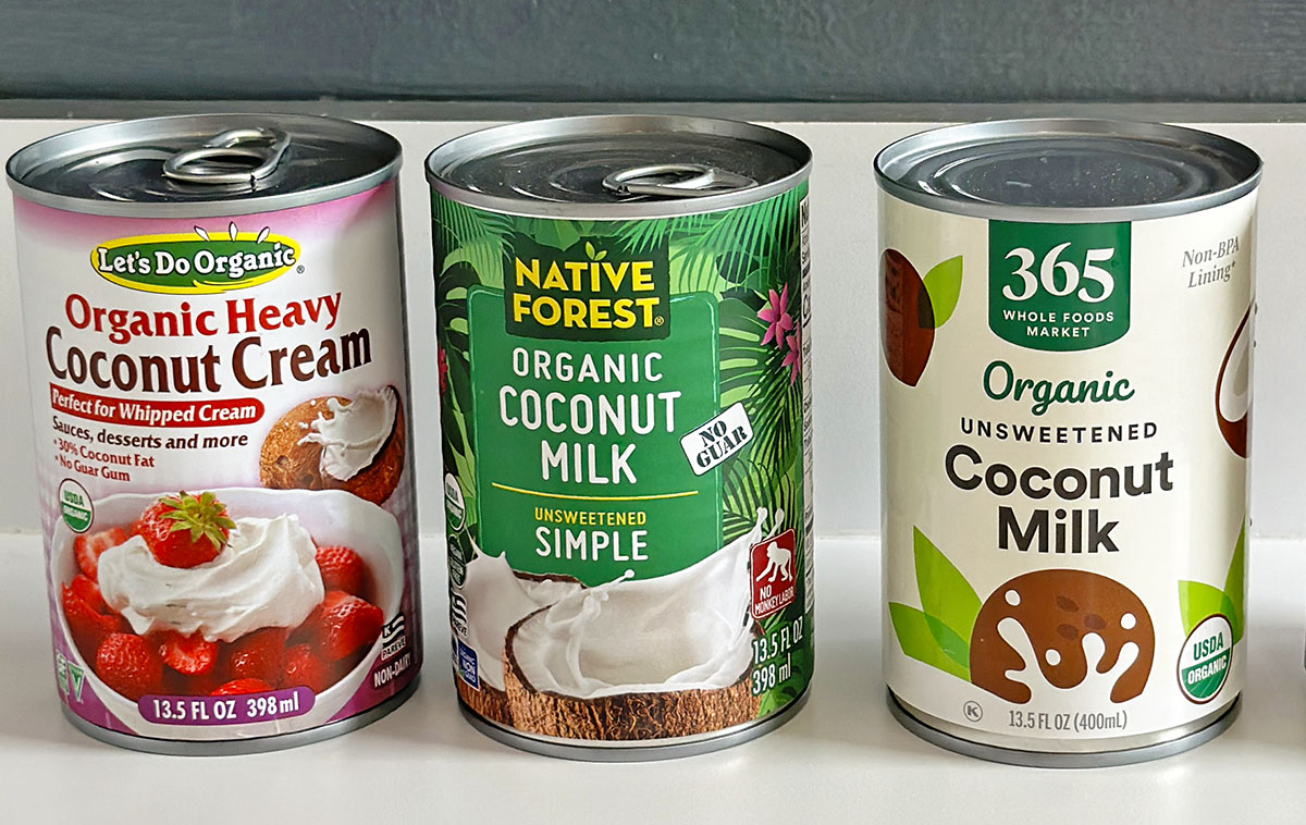 A can of coconut cream first, then a green can of coconut milk, and last a white labeled can of coconut milk on a white counter in a line.