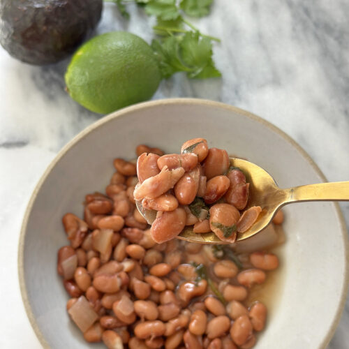 gold spoonful of beans over a gray bowl of pinto beans next to lime, avocado, and cilantro