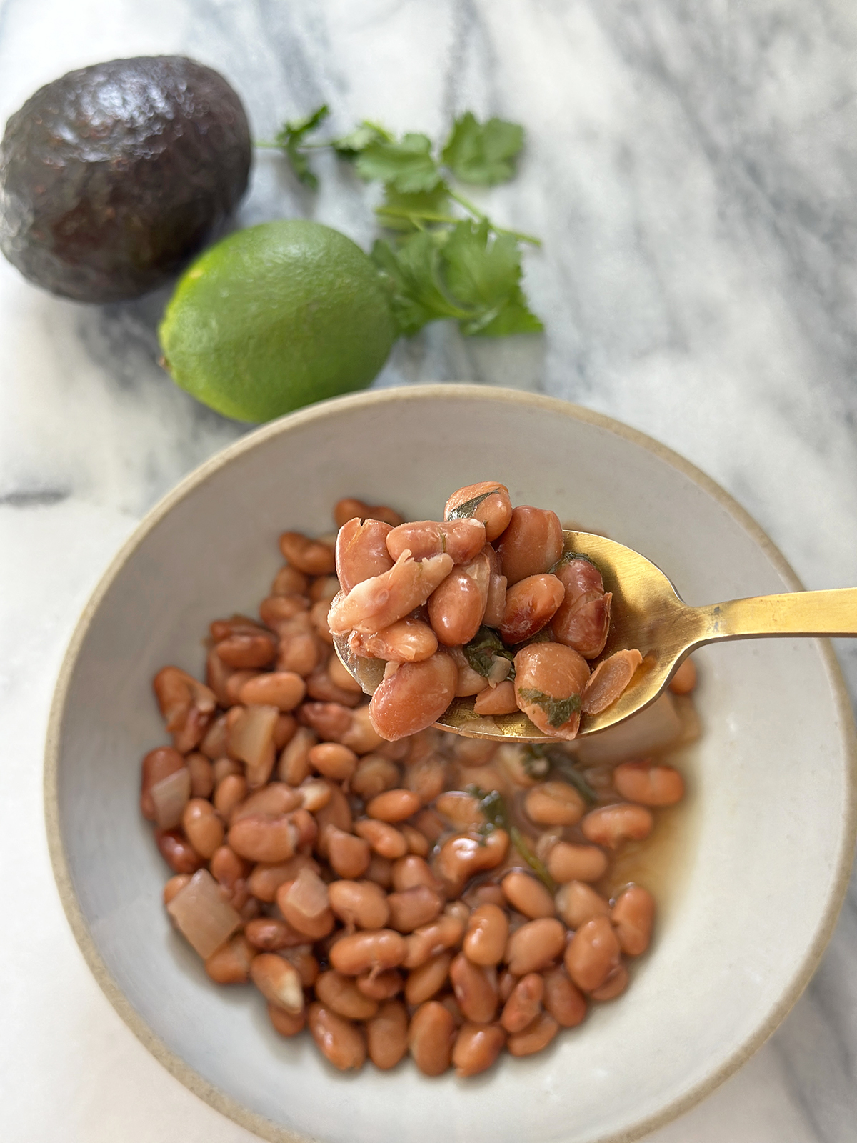 gold spoonful of beans over a gray bowl of pinto beans next to lime, avocado, and cilantro