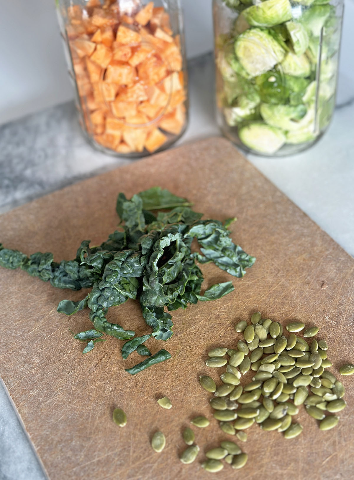 Cutting board with chopped pile of kale, pumpkin seeds, and two mason jars with sweet potatoes and Brussels sprouts chopped