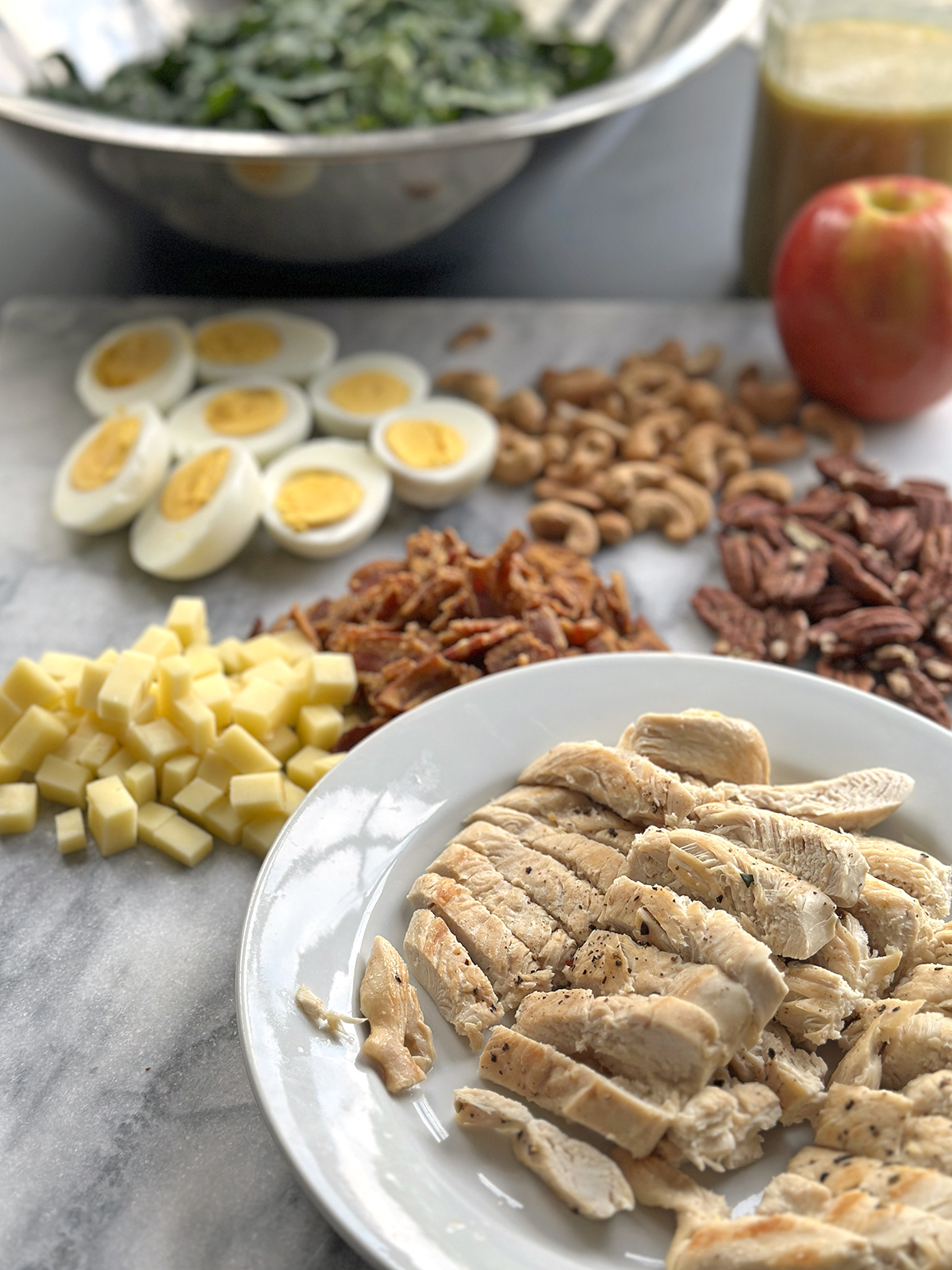 Chicken apple salad meal prep setup with roasted chicken, bacon, eggs, cheddar, nuts, and fresh kale—all ready for assembly on a white plate and marble board.