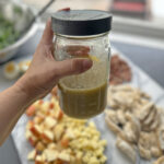 Hand holding a mason jar of homemade honey mustard vinaigrette with salad ingredients in the background