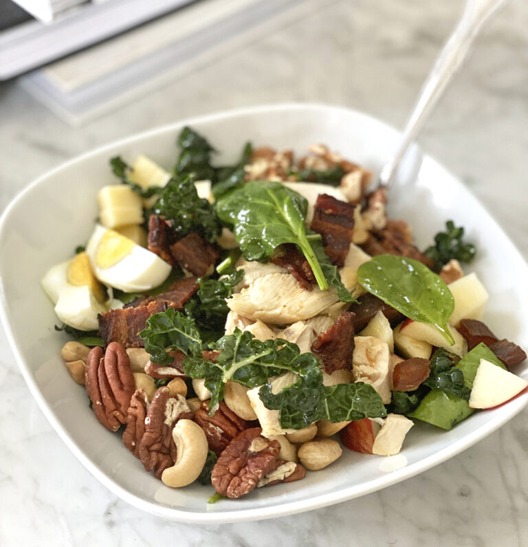 A hearty chicken apple salad with kale, spinach, roasted chicken, crispy bacon, hard-boiled egg slices, diced apples, cheddar cubes, toasted cashews, and pecans served in a white bowl.