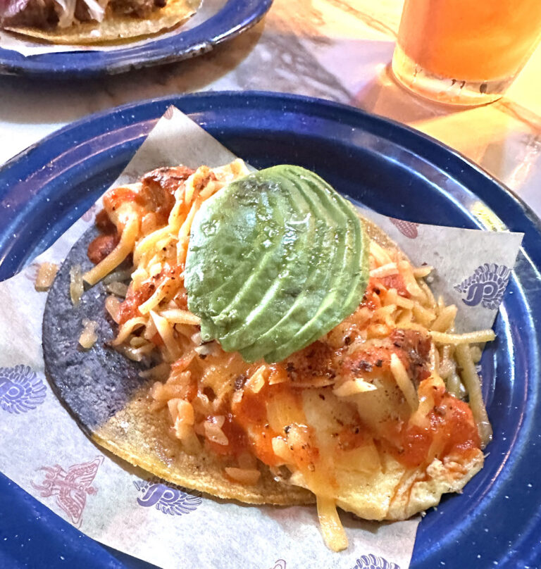 Taco on a blue and yellow corn tortilla topped with sliced avocado and cheese at Nixta Taqueria in Austin